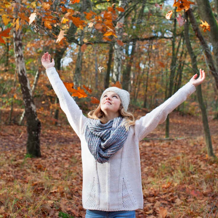 Teenage girl in forest arms raised throwing autumn leaves, looking up smiling